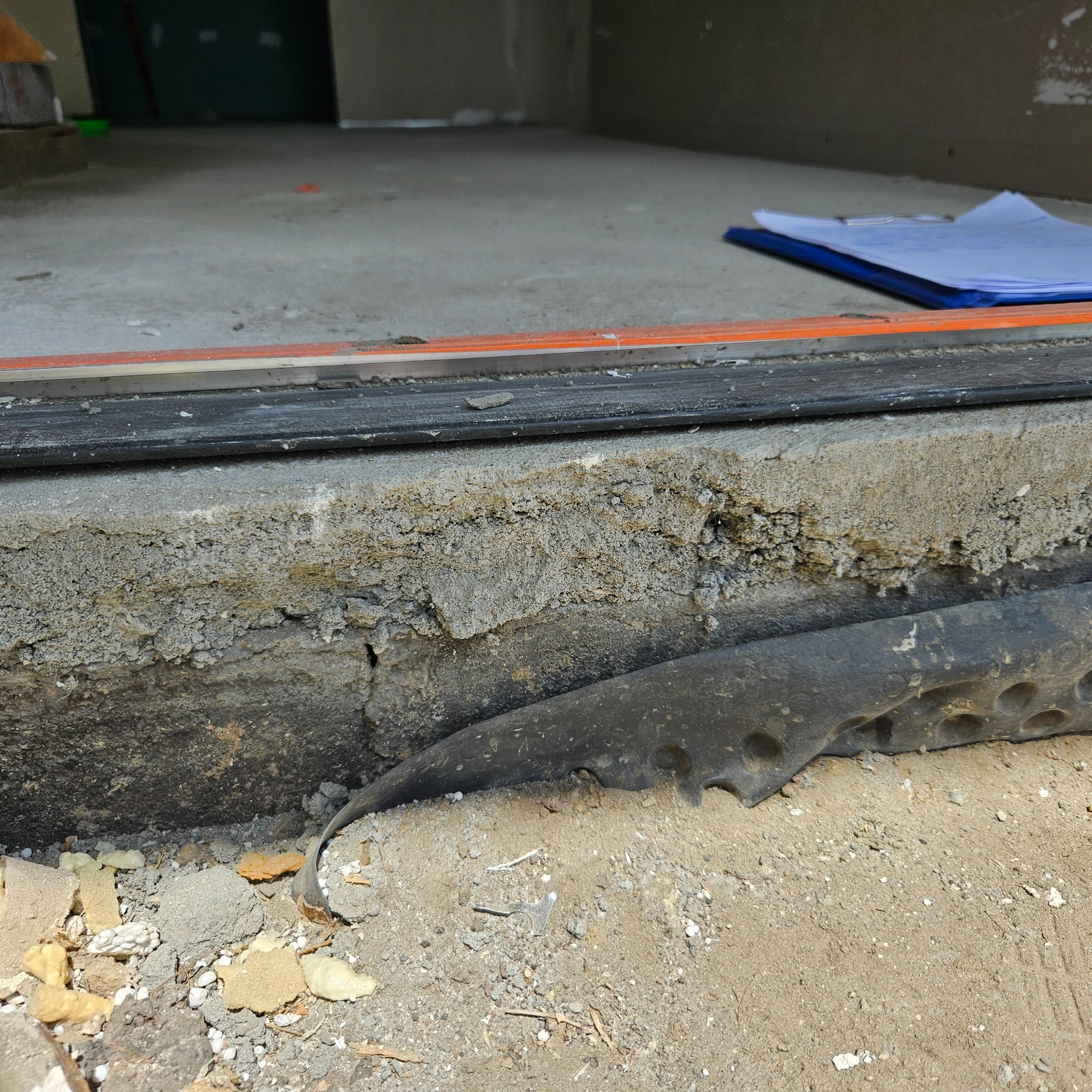 Szczegółowe ujęcie progu drzwiowego w trakcie prac wykończeniowych, widoczna warstwa izolacji, betonowa wylewka z nierówną fakturą oraz poziomica i dokumenty w tle.