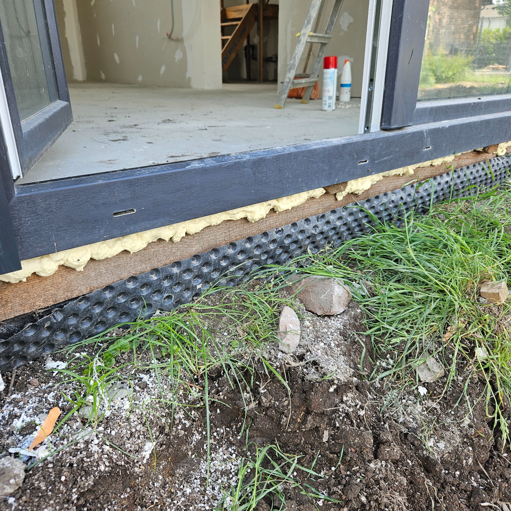 Fragment osadzania okna w nowym budynku, widoczna izolacja pianką montażową, folia kubełkowa oraz ziemia z trawą.