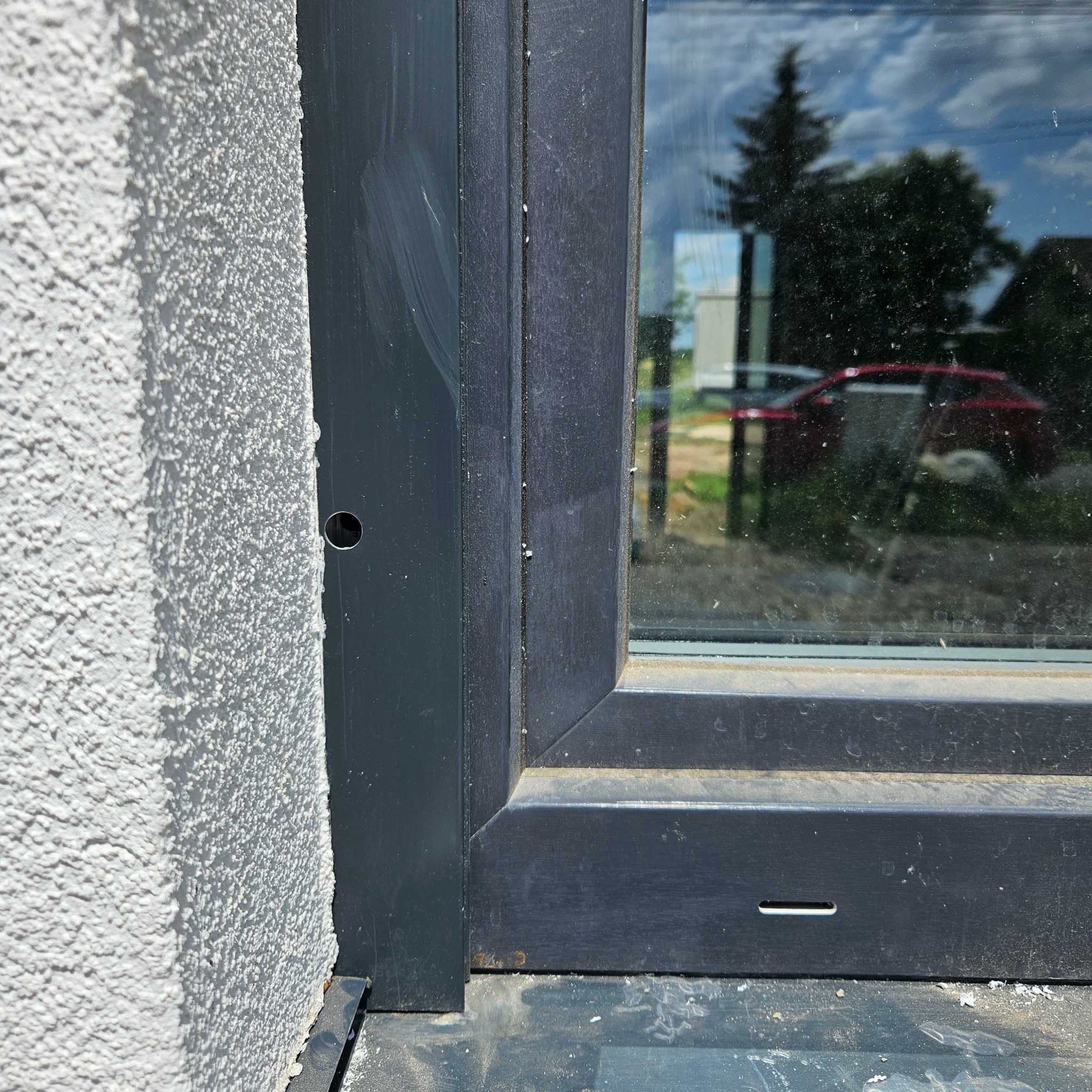 Szczegółowe ujęcie osadzonego okna PCV w szorstkiej, jasnej elewacji budynku z widocznym otworem montażowym i refleksami otoczenia na szybie.