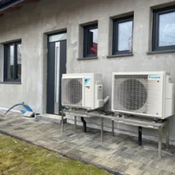 Dwa zewnętrzne agregaty klimatyzacji Daikin zamontowane na metalowych stelażach przy szarym, otynkowanym domu z grafitowymi oknami i drzwiami.