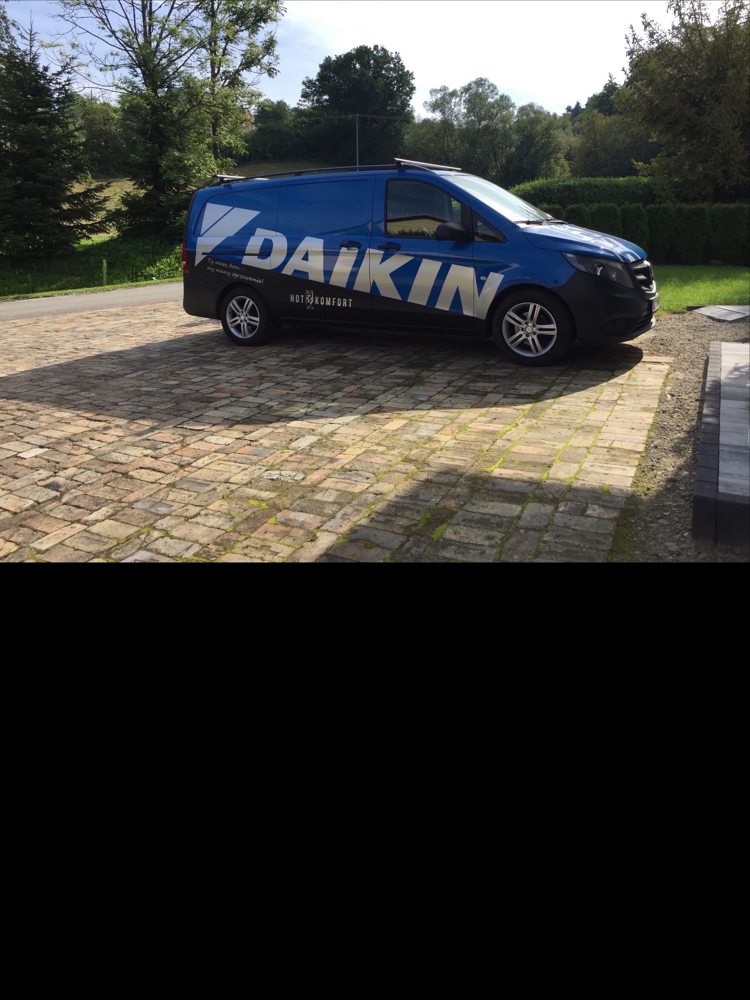 Niebieski samochód dostawczy z logo firmy na brukowanym podjeździe, w tle zieleń drzew i krzewów.