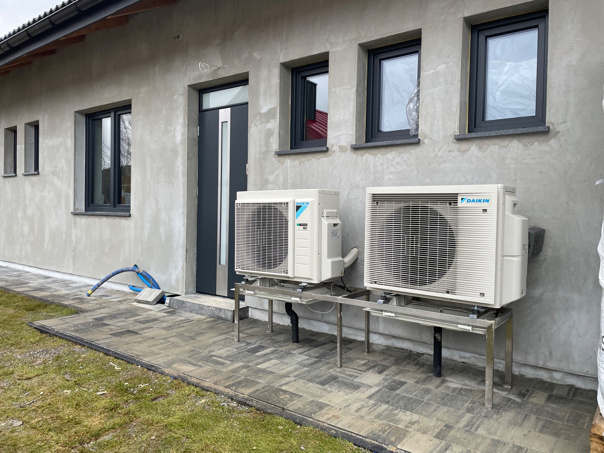 Dwa zewnętrzne agregaty klimatyzacji Daikin zamontowane na metalowych stelażach przy szarym, otynkowanym domu z grafitowymi oknami i drzwiami.