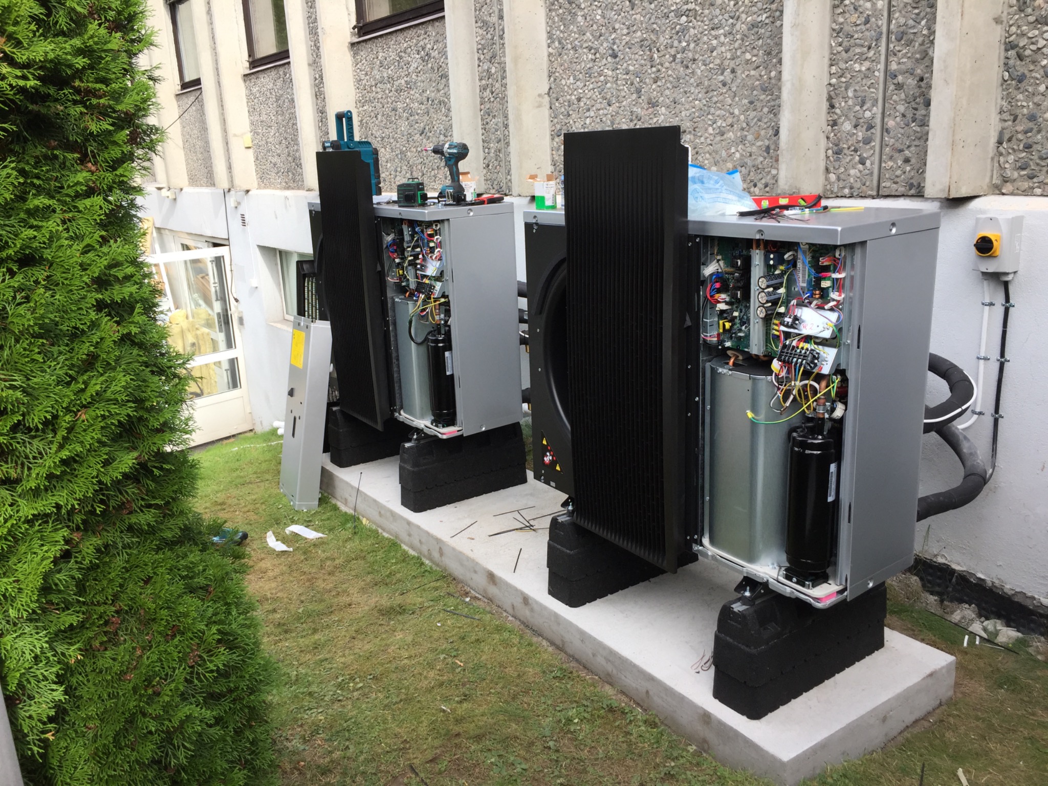 Dwa szare, otwarte panele pomp ciepła ustawione na betonowej podstawie, z widocznymi przewodami i elementami wewnętrznymi, narzędzia pracy na górze, obok zielony krzew.