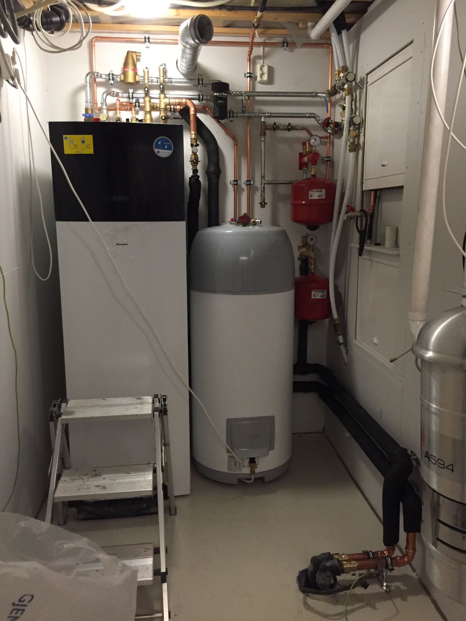 Kompletna instalacja pompy ciepła z zbiornikiem buforowym i rozbudowaną armaturą, widoczne rury miedziane, zawory i manometry w pomieszczeniu technicznym, drabina inspekcyjna obok urządzenia.