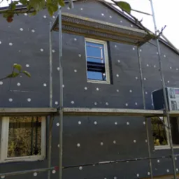 Dom w trakcie ocieplania styropianem grafitowym z widocznymi białymi kołkami mocującymi, rusztowanie metalowe przy elewacji, paczka styropianu oparta o rusztowanie.