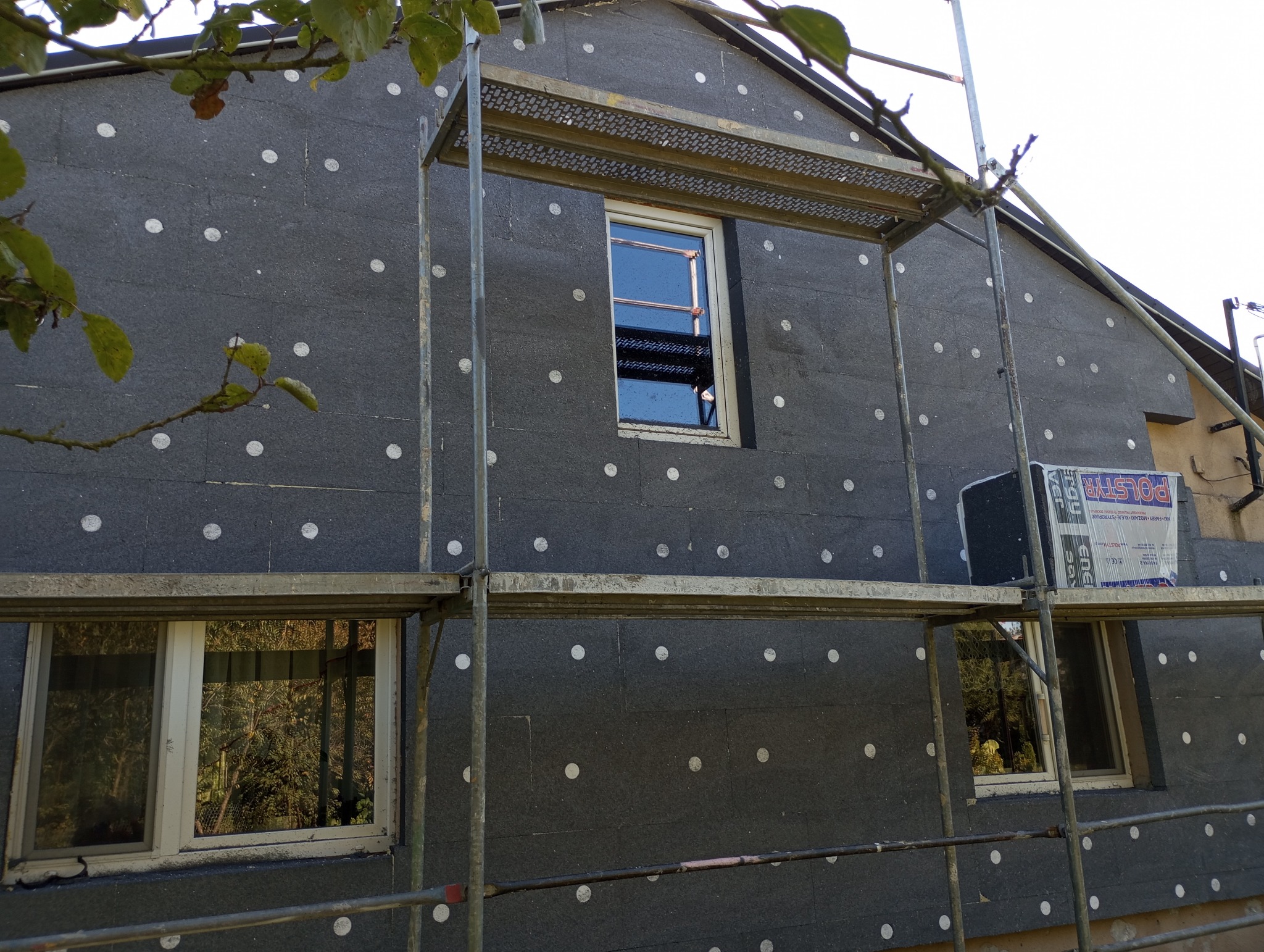 Dom w trakcie ocieplania styropianem grafitowym z widocznymi białymi kołkami mocującymi, rusztowanie metalowe przy elewacji, paczka styropianu oparta o rusztowanie.