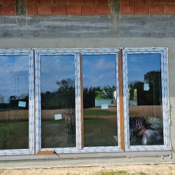 Perfekt-Mont - Czterodzielne okno z profilem Rehau w trakcie montażu w surowym stanie, widoczne cegły i beton, odblask krajobrazu w szybach.