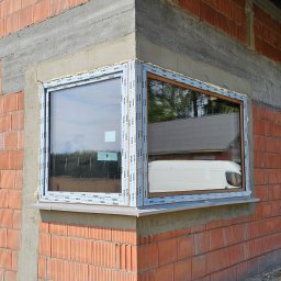 Perfekt-Mont - Narożne okno PCV z naklejkami producenta na ramie, zamontowane w surowej ścianie z czerwonej cegły ceramicznej, widoczny odblask białego samochodu dostawczego w szybie.