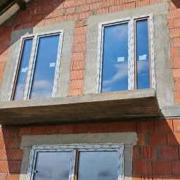 Perfekt-Mont - Montaż okien PCV w surowym murze z czerwonej cegły, widoczne pianowanie i obróbka murarska, okna marki Rehau z widocznym odbiciem nieba.