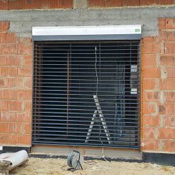 Perfekt-Mont - Montaż rolety zewnętrznej na surowej ścianie z czerwonej cegły, widoczna drabina i zwinięty kabel elektryczny.