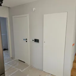 Białe drzwi wewnętrzne z czarną klamką zabezpieczoną folią, zamontowane w szarej ścianie, widoczne elementy instalacji elektrycznej i fragment schodów.