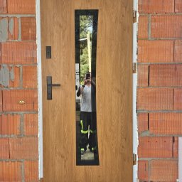 Perfekt-Mont - Brązowe drzwi wejściowe z podłużnym przeszkleniem, zamontowane w surowym murze z cegły, widoczne ślady pianki montażowej i odbicie osoby robiącej zdjęcie.