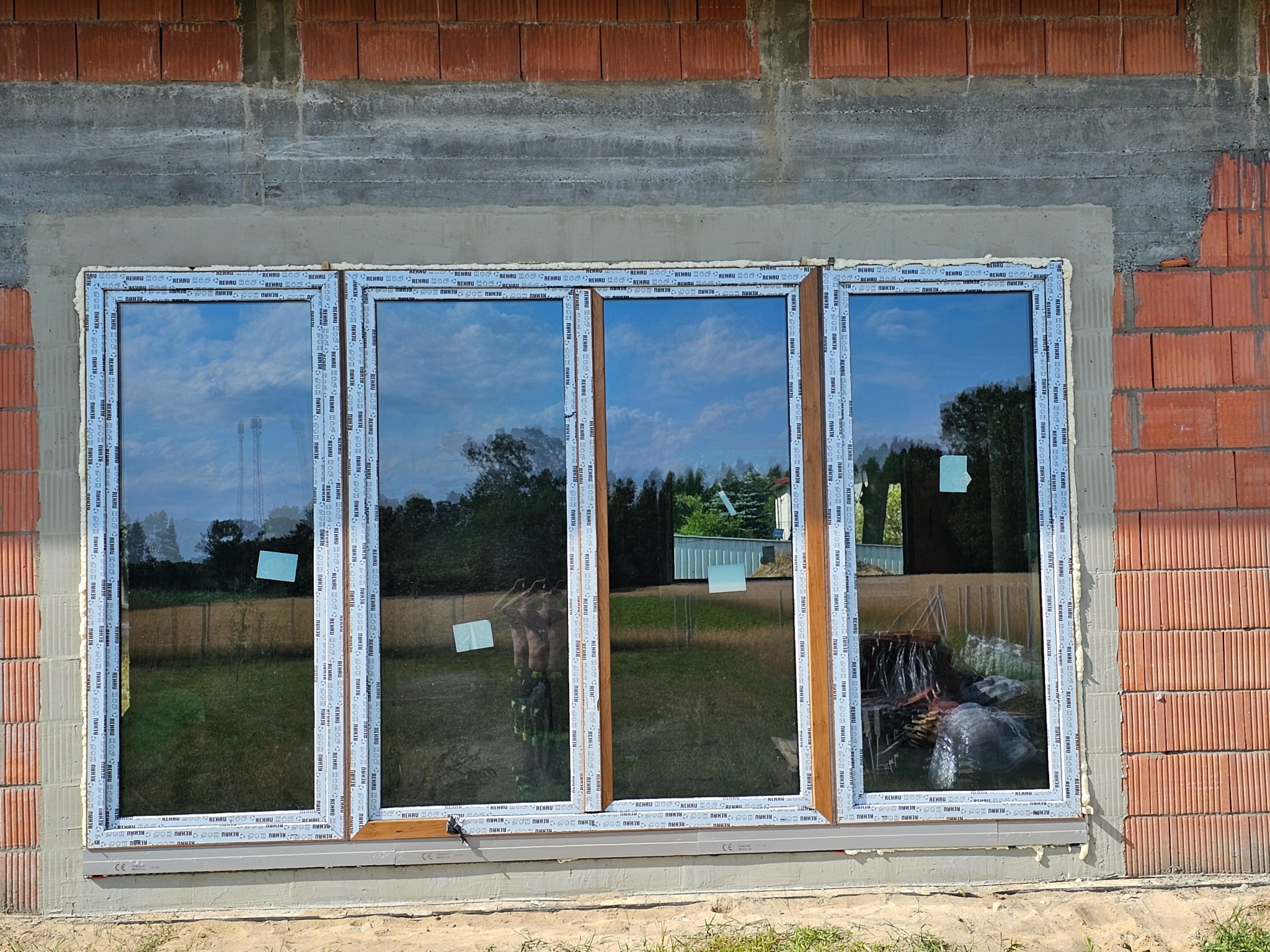 Czterodzielne okno z profilem Rehau w trakcie montażu w surowym stanie, widoczne cegły i beton, odblask krajobrazu w szybach.