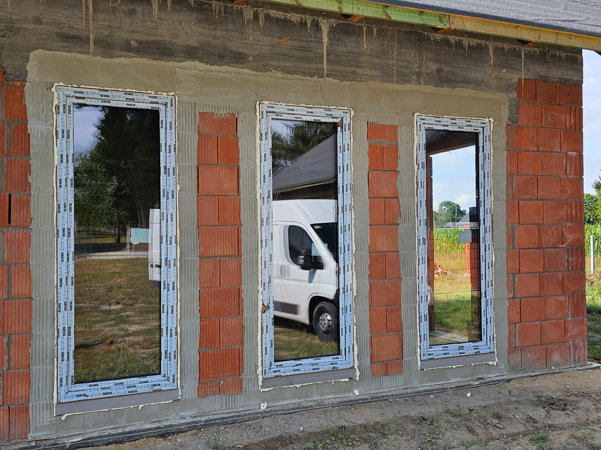 Surowy stan budynku z trzema oknami PCV marki Rehau, świeżo osadzonymi w murze z czerwonej cegły dziurawki, z pianką montażową i widocznym białym samochodem dostawczym w odbiciu szyb.