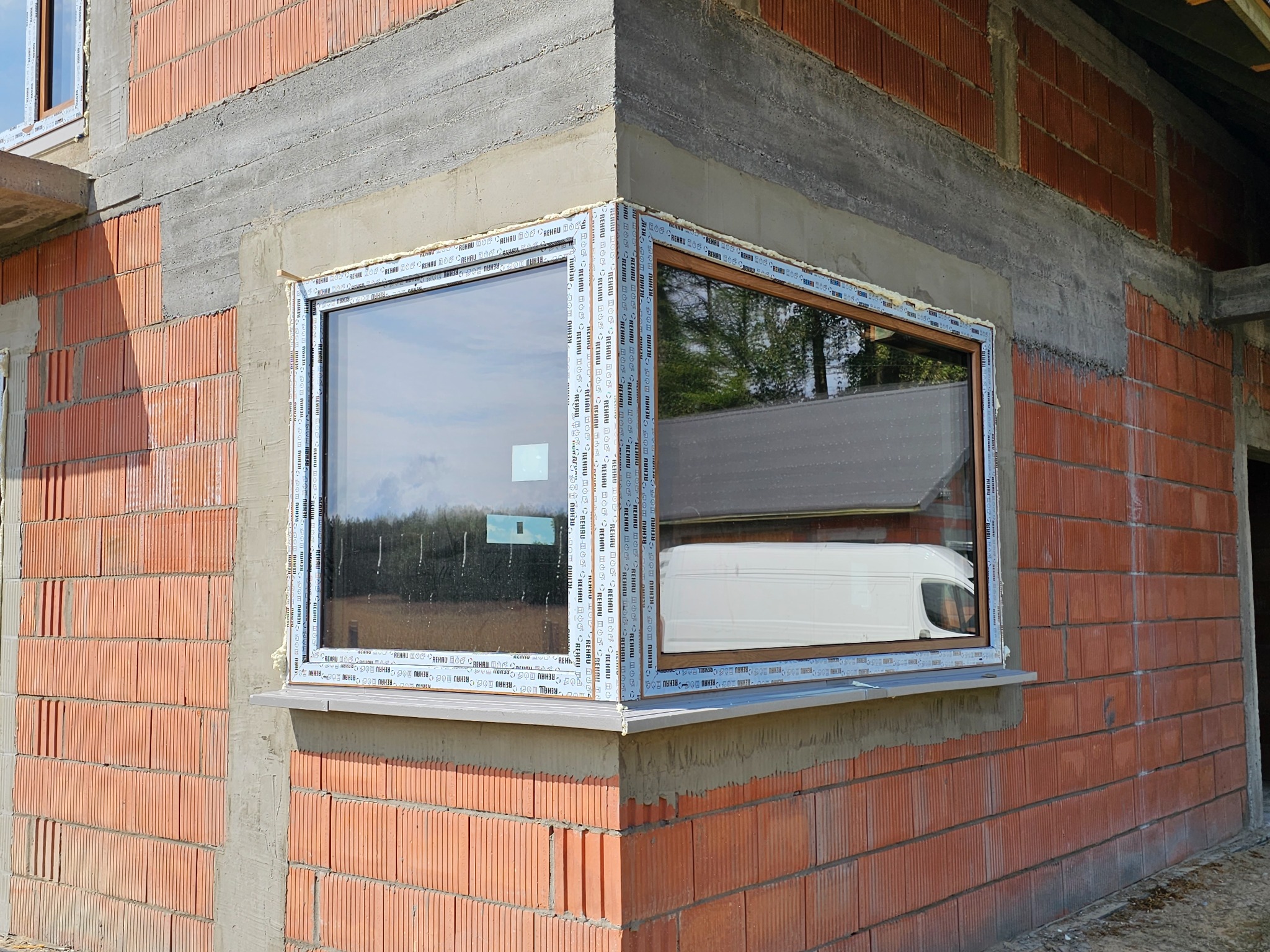 Narożne okno PCV z naklejkami producenta na ramie, zamontowane w surowej ścianie z czerwonej cegły ceramicznej, widoczny odblask białego samochodu dostawczego w szybie.