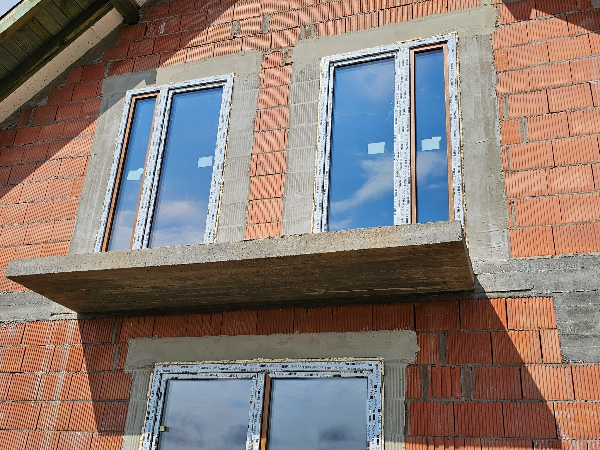 Montaż okien PCV w surowym murze z czerwonej cegły, widoczne pianowanie i obróbka murarska, okna marki Rehau z widocznym odbiciem nieba.