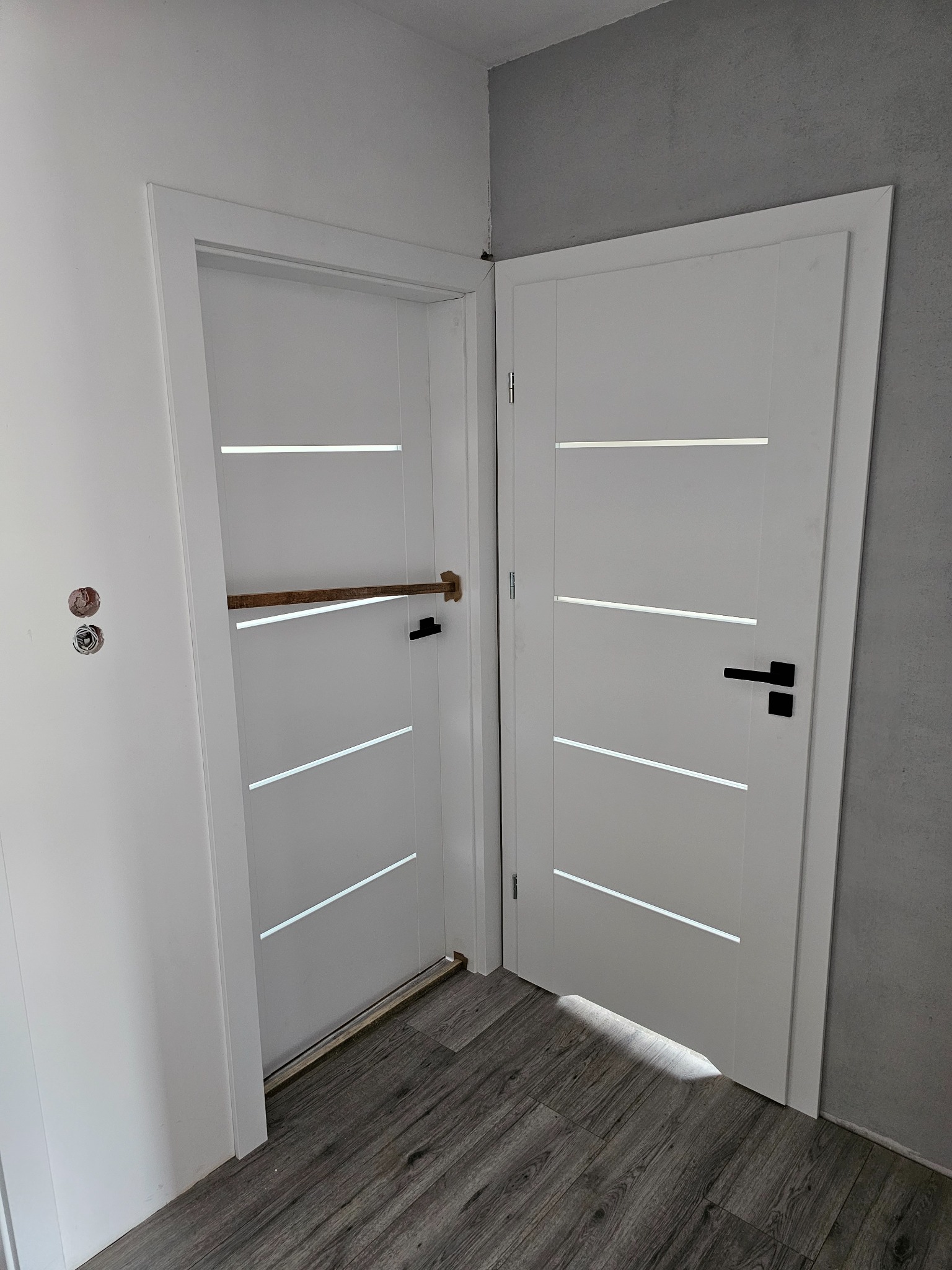 Białe, nowoczesne drzwi wewnętrzne z poziomymi przeszkleniami, jedne zamontowane i otwarte z drewnianą rozpórką, drugie z czarną klamką, widoczne otwory po starych zamkach, podłoga imitująca drewno.