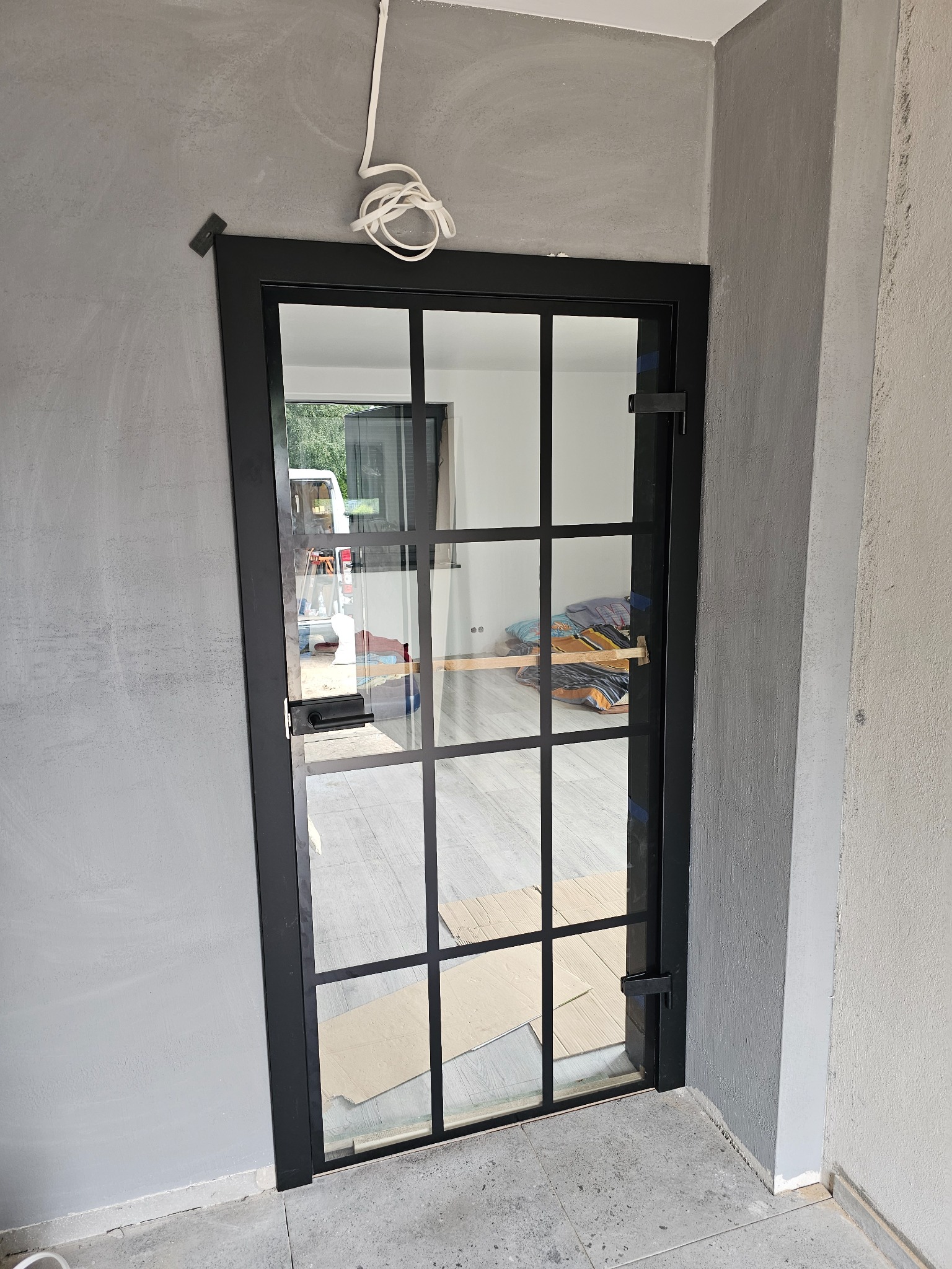 Czarna, przeszklona drzwi wewnętrzne z metalowymi szprosami, widok na wnętrze z pościelą i częściowo widocznym samochodem dostawczym za oknem, szare ściany w surowym wykończeniu.