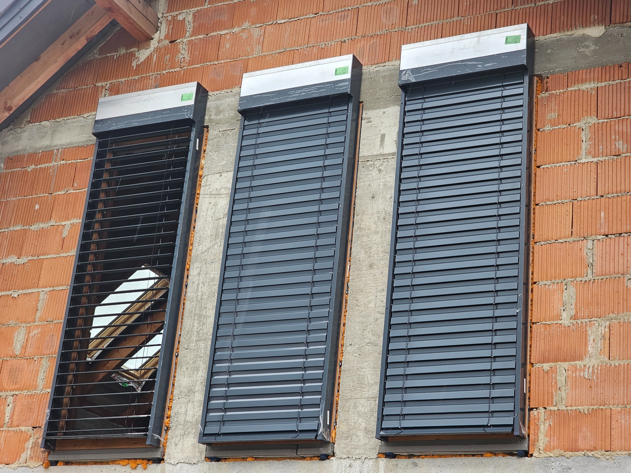 Trzy okna dachowe z zamontowanymi szarymi roletami zewnętrznymi, z których jedna jest otwarta, ukazując okno dachowe i konstrukcję dachu, na tle muru z czerwonej cegły.