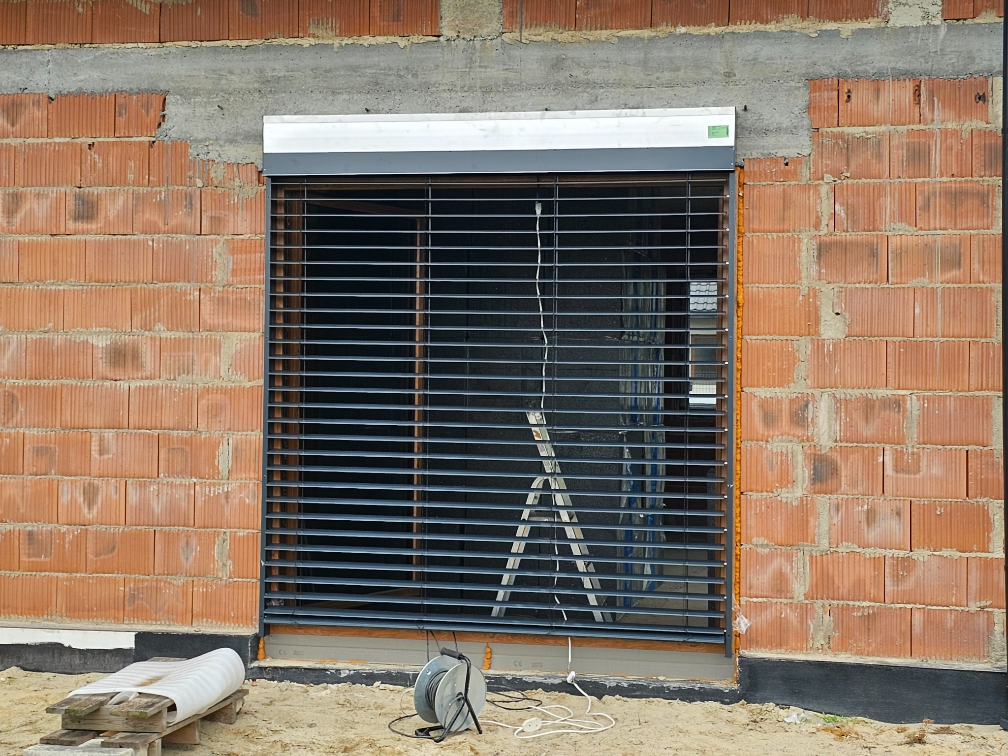 Montaż rolety zewnętrznej na surowej ścianie z czerwonej cegły, widoczna drabina i zwinięty kabel elektryczny.