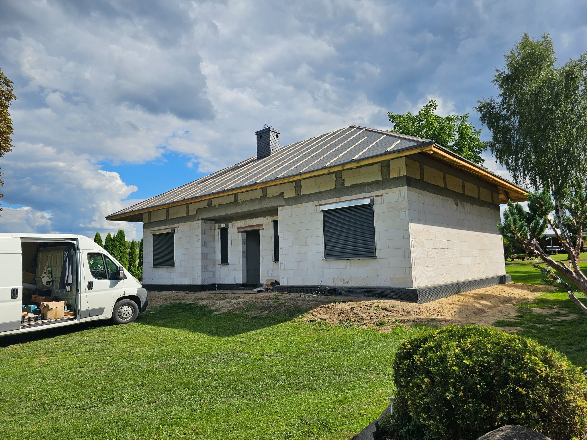 Surowy stan domu jednorodzinnego z szarej cegły, częściowo zadaszony, z zamontowanymi roletami zewnętrznymi w oknach, widoczny bus serwisowy z otwartymi drzwiami.