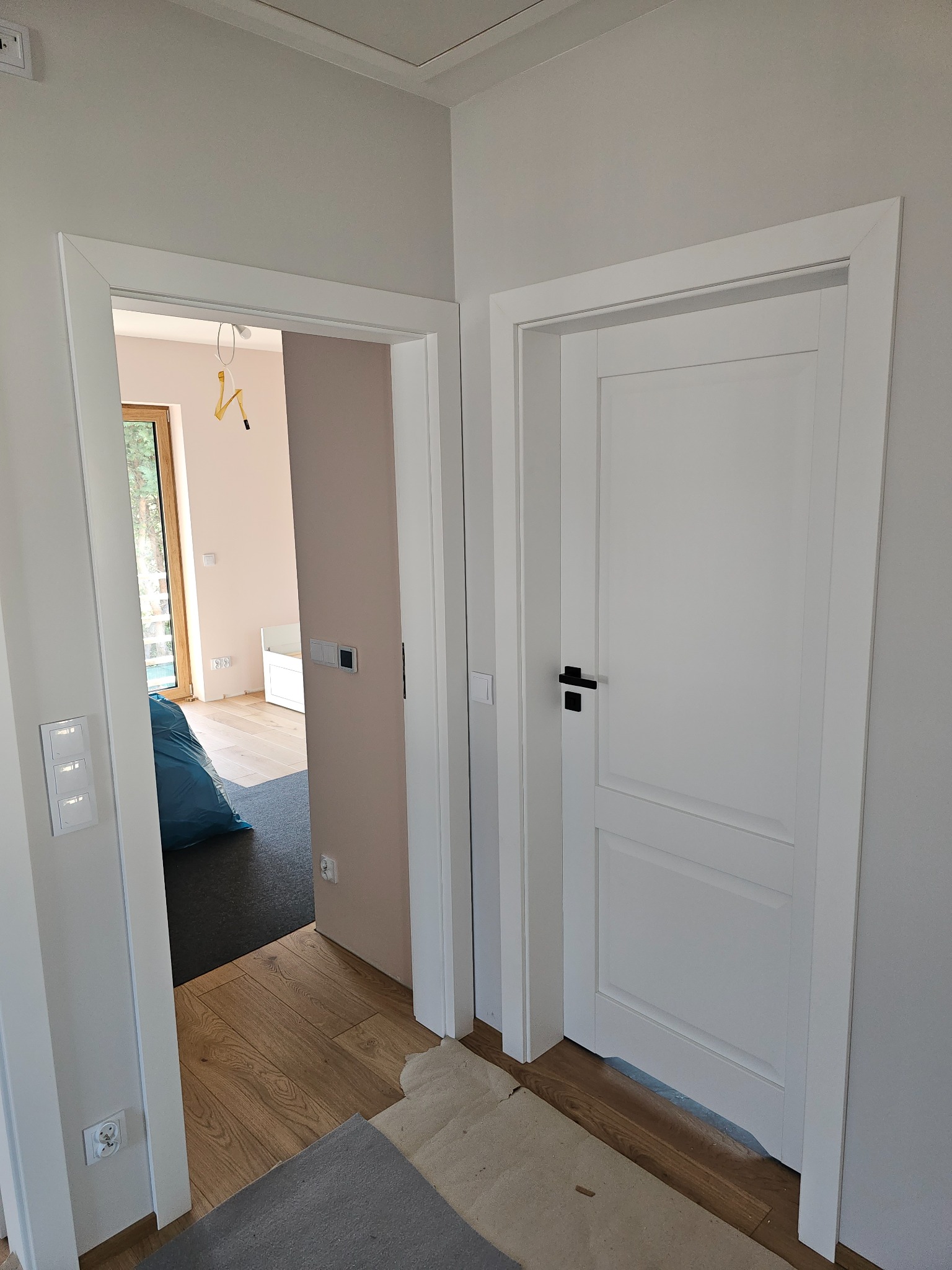 Nowoczesne, białe drzwi wewnętrzne z czarną klamką w minimalistycznym wnętrzu, widoczne przez otwarte drzwi, w tle pokój w trakcie wykańczania.