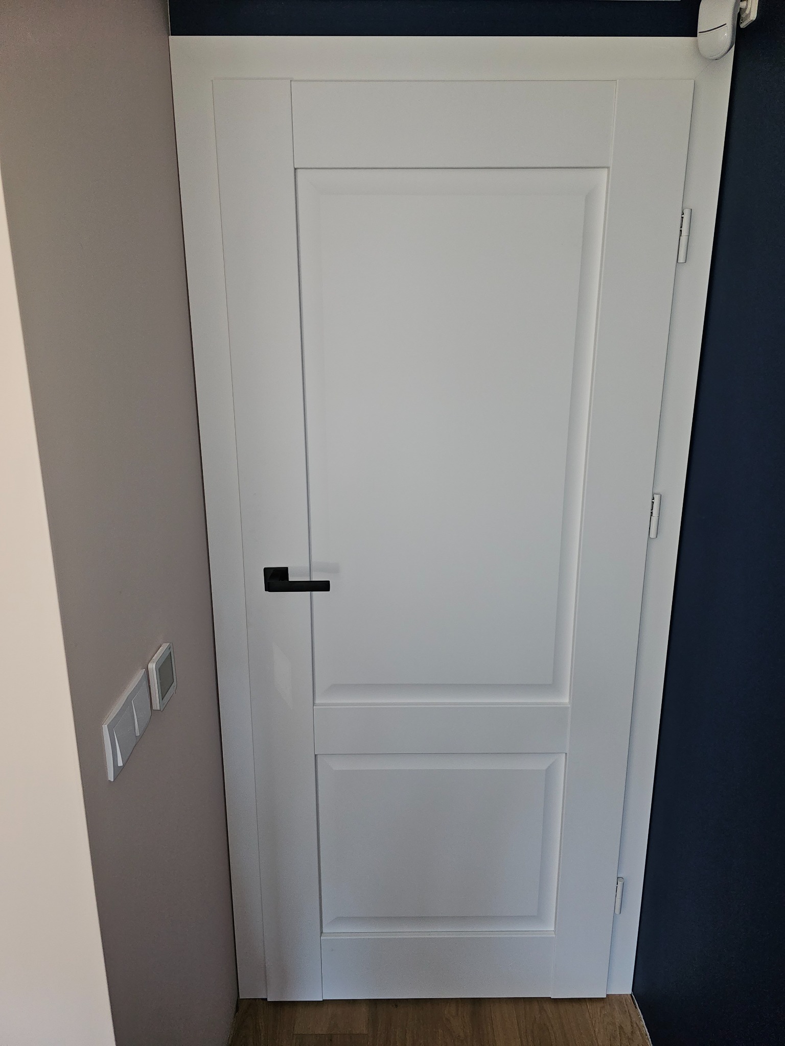 Białe drzwi wewnętrzne z czarną klamką na tle szarej i granatowej ściany, widoczne włączniki światła i drewniana podłoga.