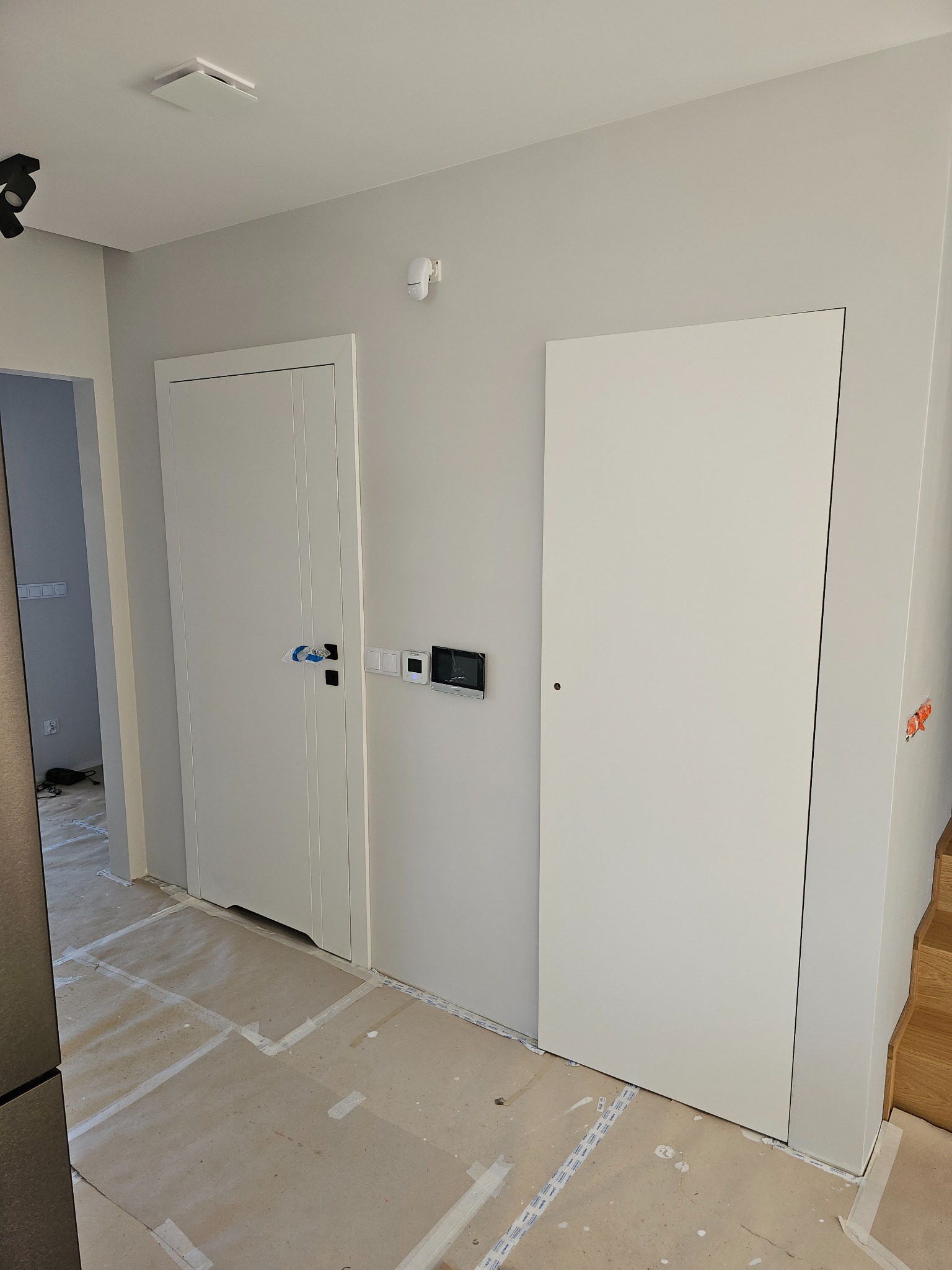 Białe drzwi wewnętrzne z czarną klamką zabezpieczoną folią, zamontowane w szarej ścianie, widoczne elementy instalacji elektrycznej i fragment schodów.