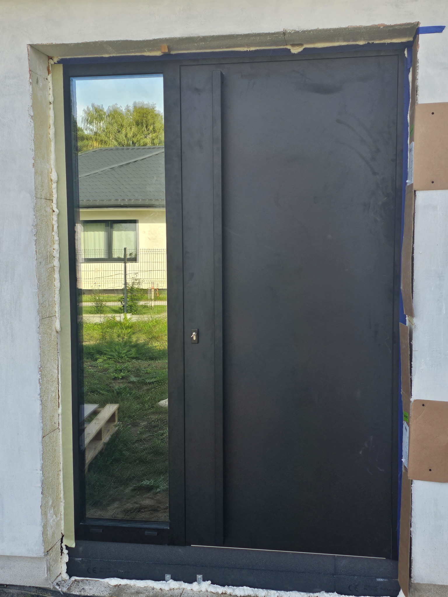 Ciemne drzwi wejściowe z bocznym przeszkleniem, widoczne pianka montażowa i taśma zabezpieczająca na ścianie, w przeszkleniu odbicie domu i zieleni.