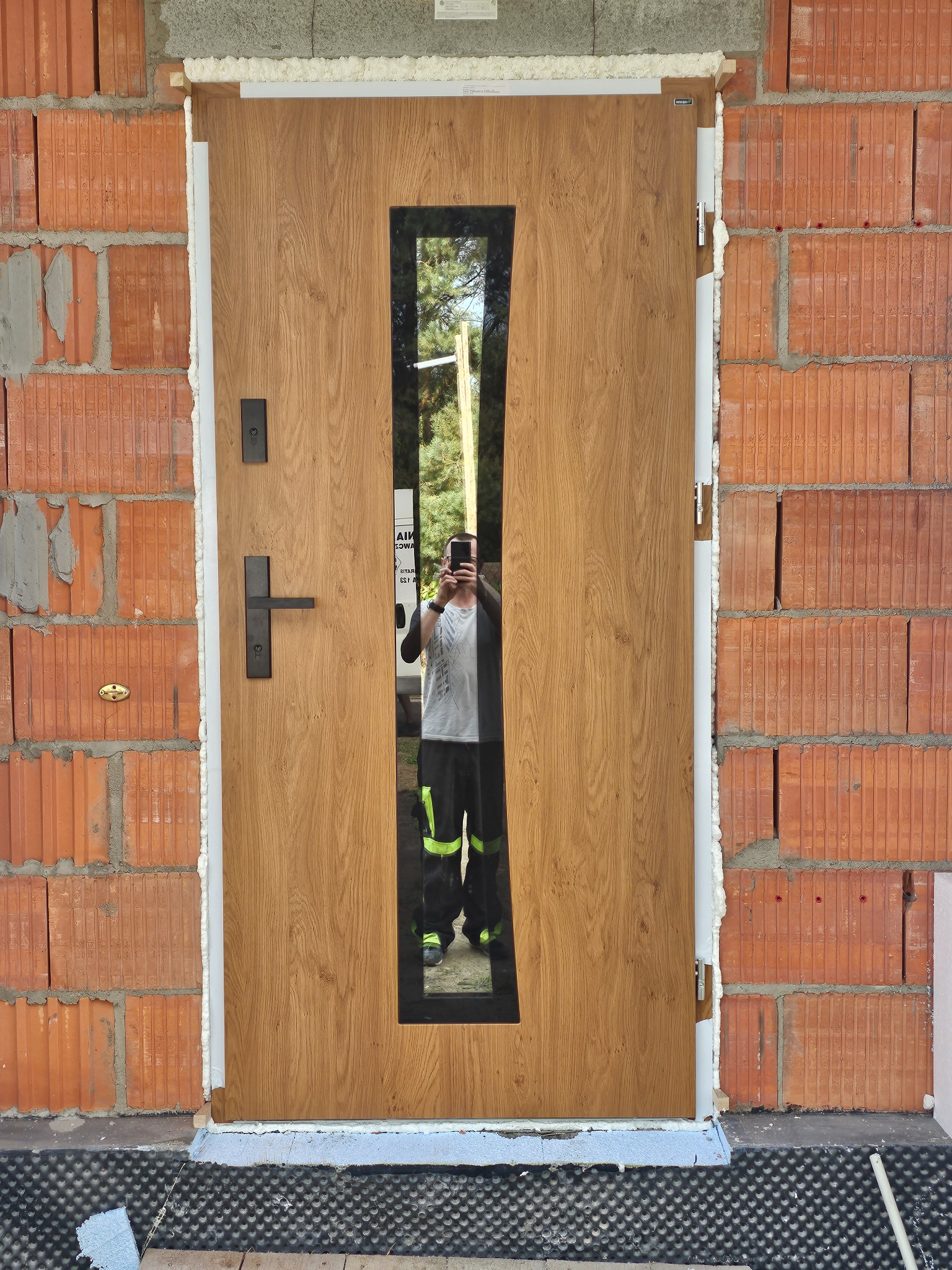 Brązowe drzwi wejściowe z podłużnym przeszkleniem, zamontowane w surowym murze z cegły, widoczne ślady pianki montażowej i odbicie osoby robiącej zdjęcie.