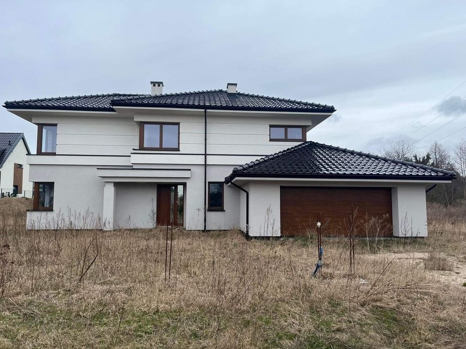 Nowoczesny, dwukondygnacyjny dom z garażem, wykończony w białym tynku z ciemną dachówką, widoczny w pochmurny dzień na tle trawiastej działki.