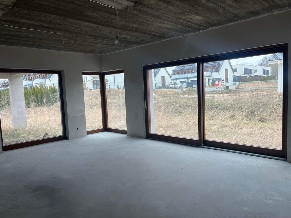 Surowy stan wnętrza domu murowanego z dużymi oknami, widok na zewnątrz na sąsiednie domy i niezagospodarowany teren, betonowa podłoga i sufit, niedokończone ściany.