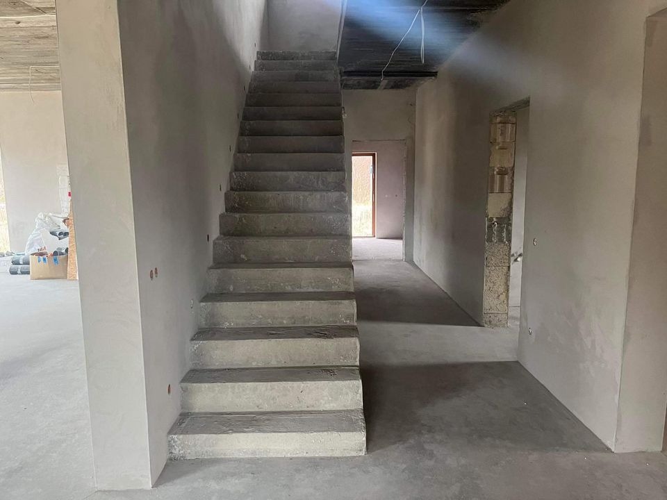 Wnętrze w stanie surowym: betonowe schody prowadzą na wyższą kondygnację, ściany otynkowane na szaro, surowy strop i widoczne instalacje.