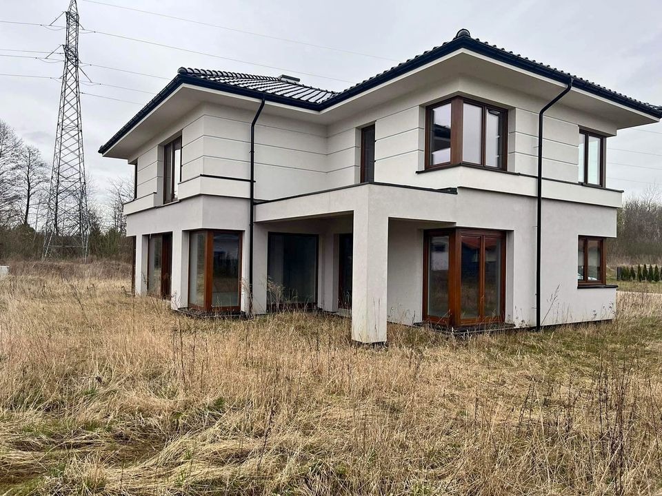 Dwukondygnacyjny, nowoczesny dom z płaskim dachem, białymi ścianami i ciemnymi ramami okiennymi, otoczony suchą trawą, z widoczną wieżą wysokiego napięcia w tle.