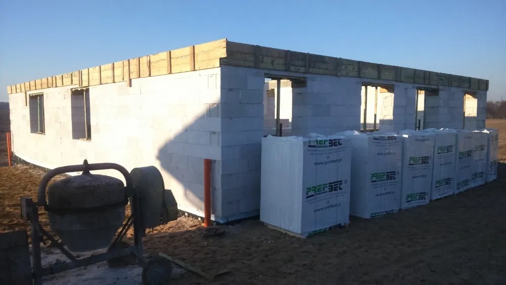 Surowy stan budynku z białych bloczków, widoczne otwory okienne i drzwiowe, drewniane nadproża, obok składowane palety z materiałem budowlanym, w tle krajobraz polny, na pierwszym planie betoniarka.