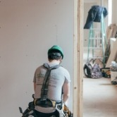Pracownik w zielonym kasku, z tyłu, podczas szpachlowania ściany w jasnym pomieszczeniu z widoczną drabiną i torbą w tle.