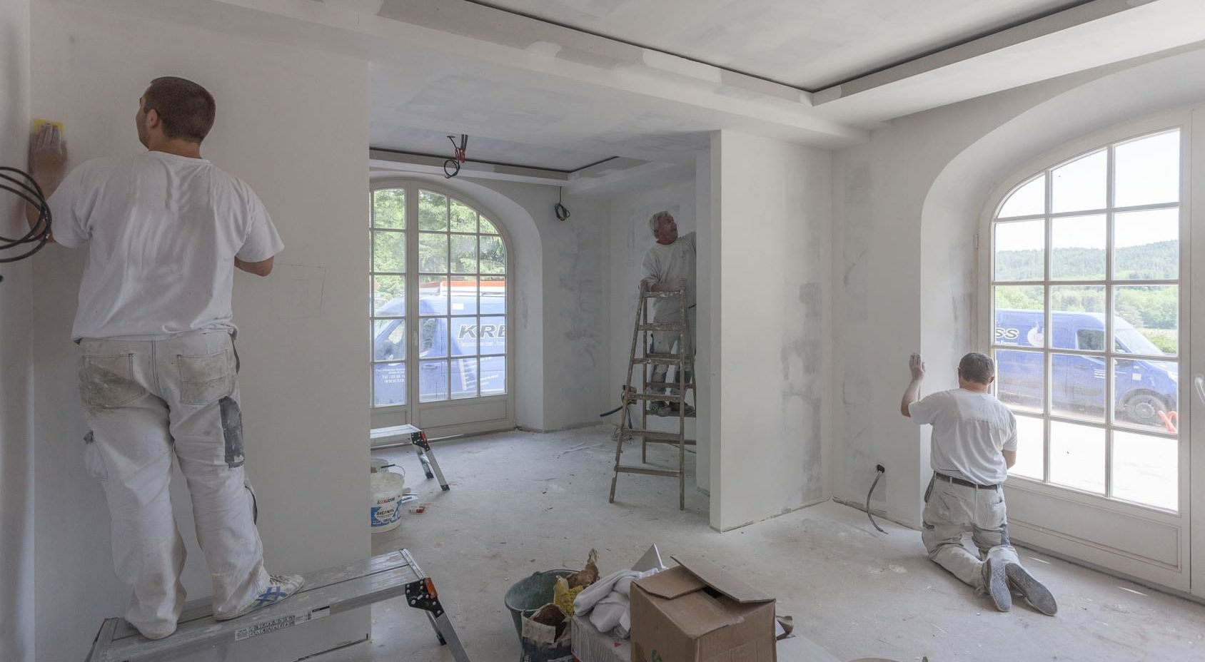 Wnętrze w trakcie przygotowania do malowania: pracownicy w białych kombinezonach szlifują ściany i sufit, widoczne drabiny, szpachle, okna łukowe z widokiem na zewnątrz i firmowy samochód dostawczy.