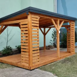 Drewniana altana z płaskim, ciemnym dachem, z elementami dekoracyjnymi z listew, umieszczona na trawniku przed budynkiem z dużymi oknami.