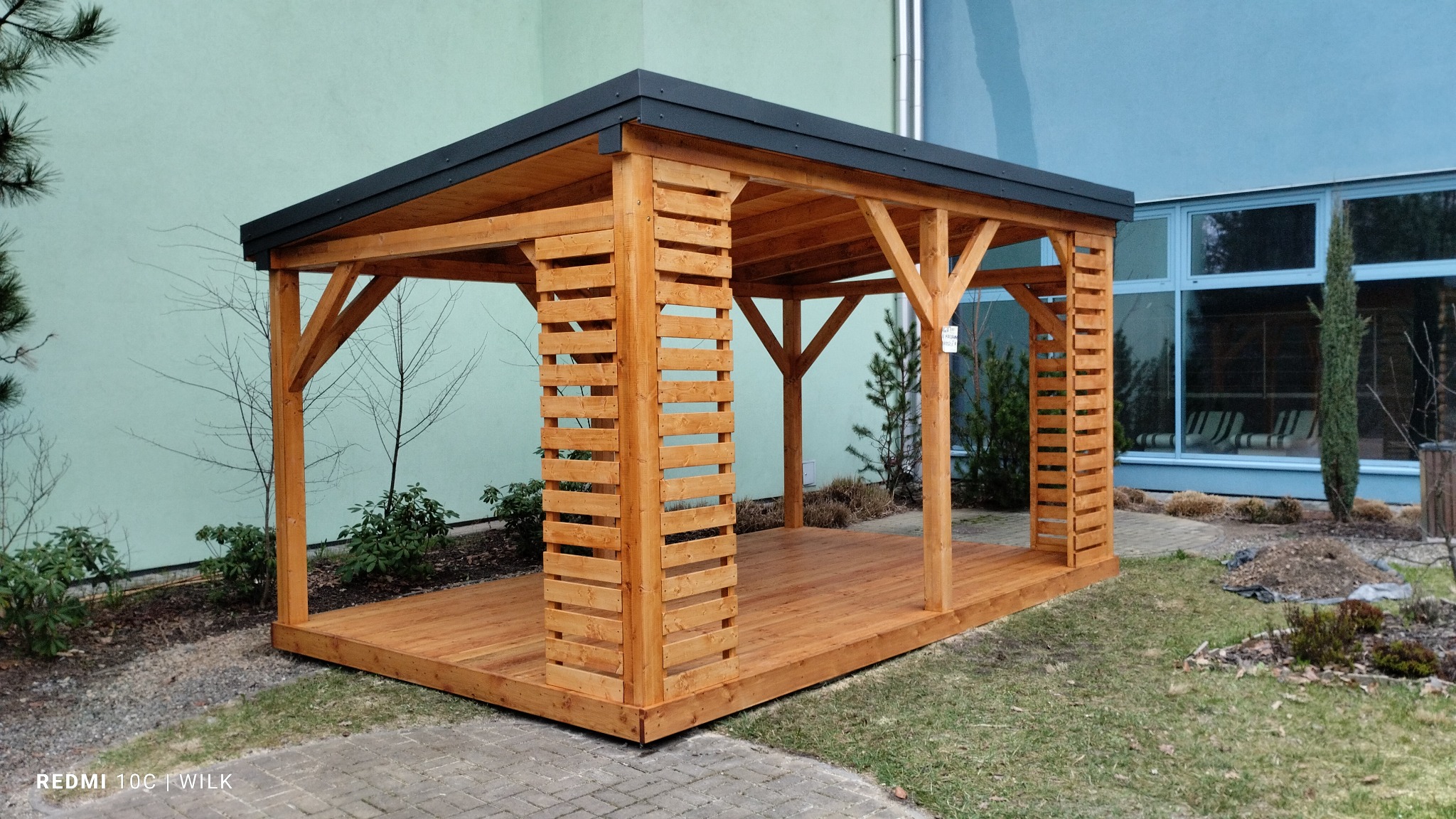 Drewniana altana z płaskim, ciemnym dachem, z elementami dekoracyjnymi z listew, umieszczona na trawniku przed budynkiem z dużymi oknami.