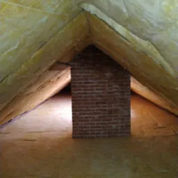 Poddasze z izolacją z wełny mineralnej w kolorze żółtym, widoczny komin z czerwonej cegły. Izolacja przymocowana sznurkami do krokwi.