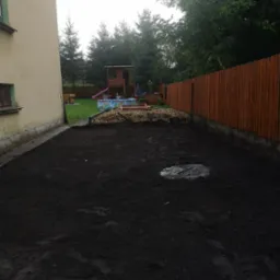 Przygotowany teren pod trawnik z widoczną świeżo rozplantowaną czarną ziemią, w tle drewniany płot i elementy placu zabaw.