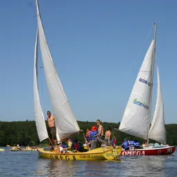 Dwie żaglówki Omega na jeziorze, jedna żółta z grupą dzieci w kamizelkach ratunkowych i dorosłym, druga czerwona z napisem 'Szarlota', w tle las i kajaki.