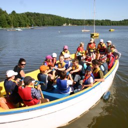 PIOTR TROJANOWSKI CRS AQUA - Grupa dzieci i dorosłych w łodzi wiosłowej, wszyscy w kamizelkach ratunkowych, na jeziorze z żaglówkami w tle.