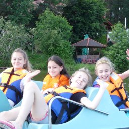 PIOTR TROJANOWSKI CRS AQUA - Grupa uśmiechniętych dziewcząt w kamizelkach ratunkowych macha rękami, siedząc na turkusowym rowerze wodnym na jeziorze, w tle widoczny las i altana.