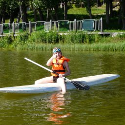 PIOTR TROJANOWSKI CRS AQUA - Dziewczyna w pomarańczowej kamizelce asekuracyjnej i niebieskich okularach pływackich siedzi na desce SUP na jeziorze, trzymając wiosło i pokazując kciuk w górę.