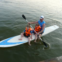 PIOTR TROJANOWSKI CRS AQUA - Mężczyzna wiosłuje na desce SUP z dwójką dzieci w pomarańczowych kamizelkach ratunkowych na spokojnej wodzie.