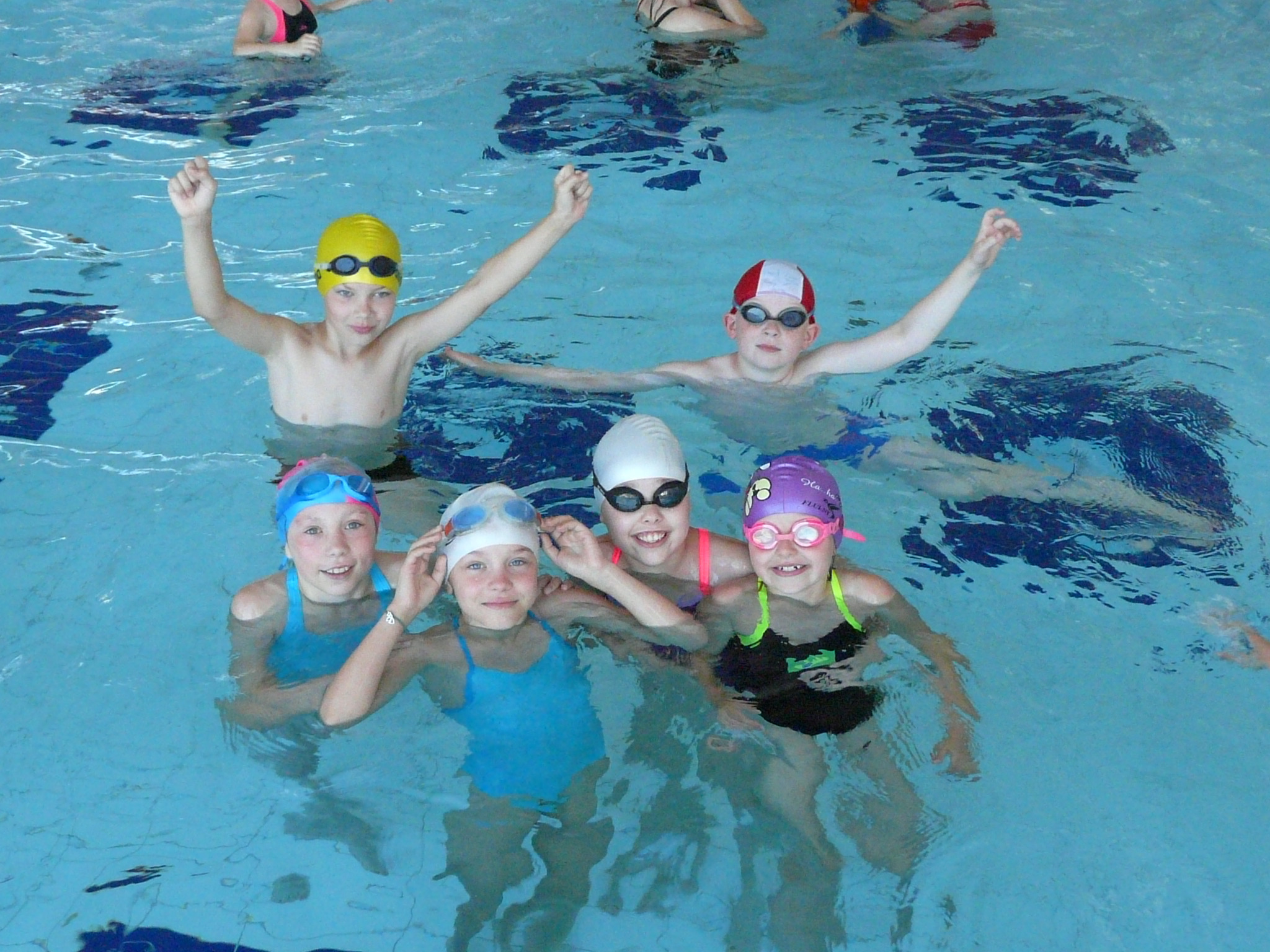 Grupa dzieci w czepkach i okularach pływackich, unosząca się w basenie z jasnoniebieską wodą, uśmiechnięte twarze skierowane w stronę kamery, dwoje dzieci z podniesionymi rękami.
