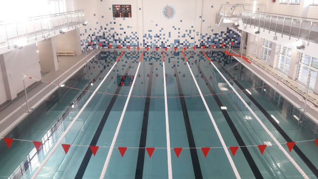 Widok z góry na pusty basen sportowy z sześcioma torami, oddzielonymi linami z czerwonymi flagami, na ścianie mozaika z niebieskich i białych płytek, na górze balkon.