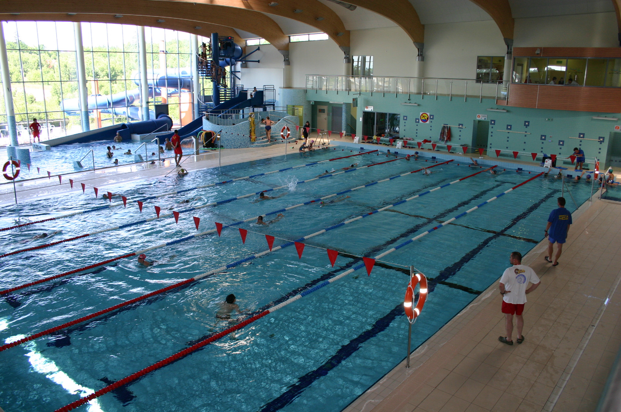 Wnętrze krytej pływalni z torami, ratownikami i zjeżdżalnią w tle; widoczne osoby pływające i bawiące się w wodzie.