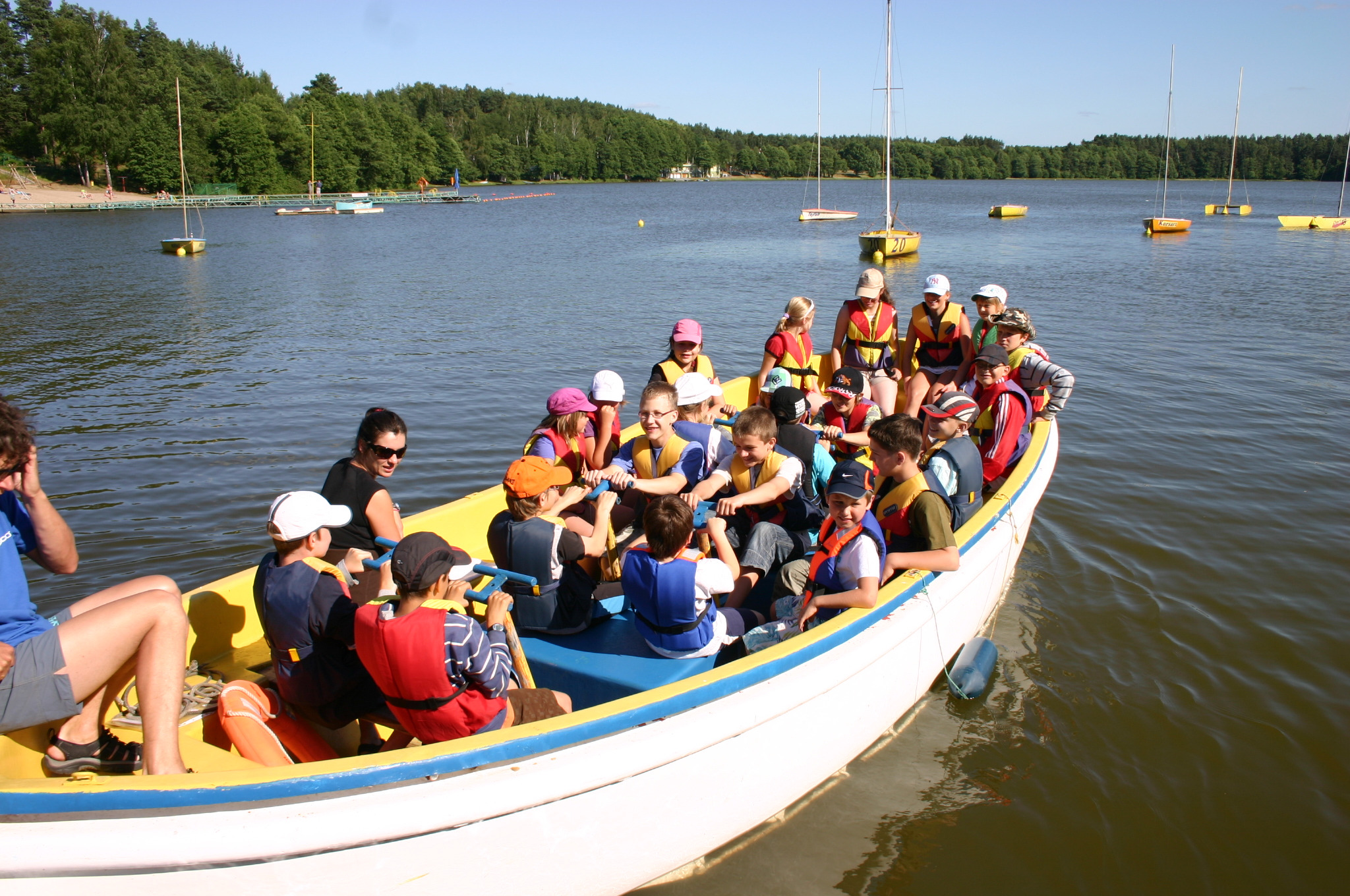 Grupa dzieci i dorosłych w łodzi wiosłowej, wszyscy w kamizelkach ratunkowych, na jeziorze z żaglówkami w tle.